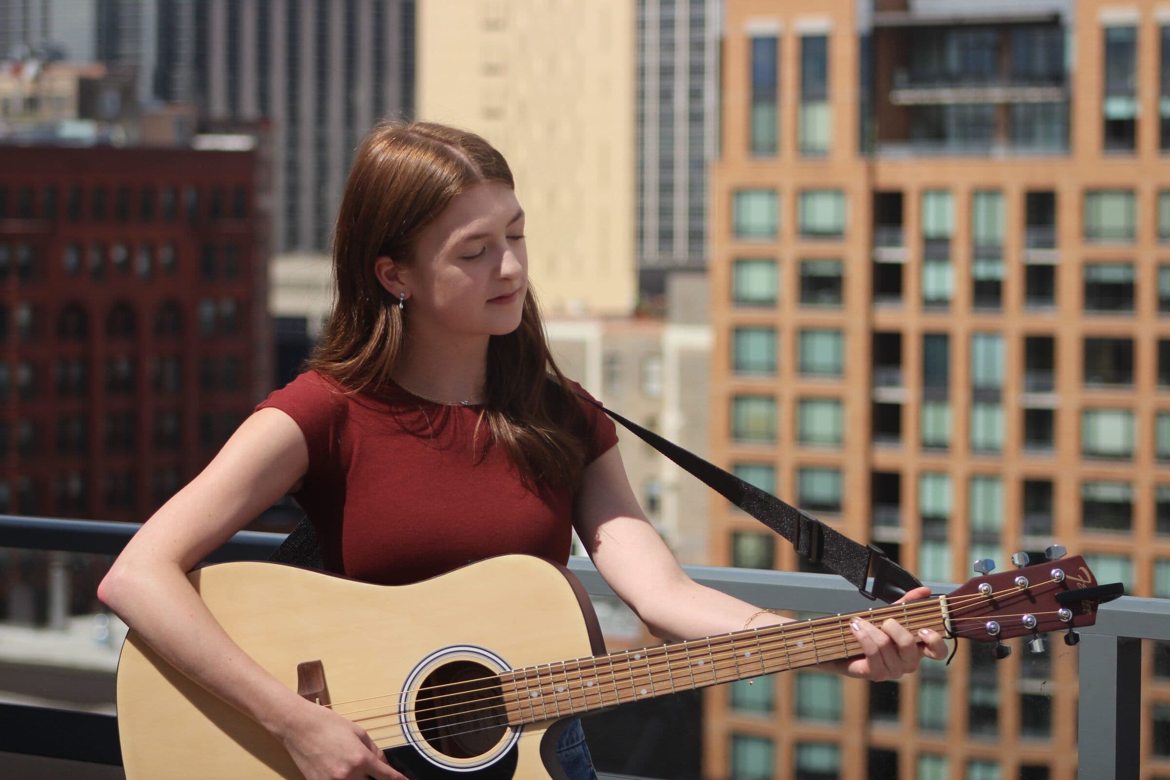 Thelma plays acoustic guitar on a rooftop, eyes closed, the Chicago skyline behind her.