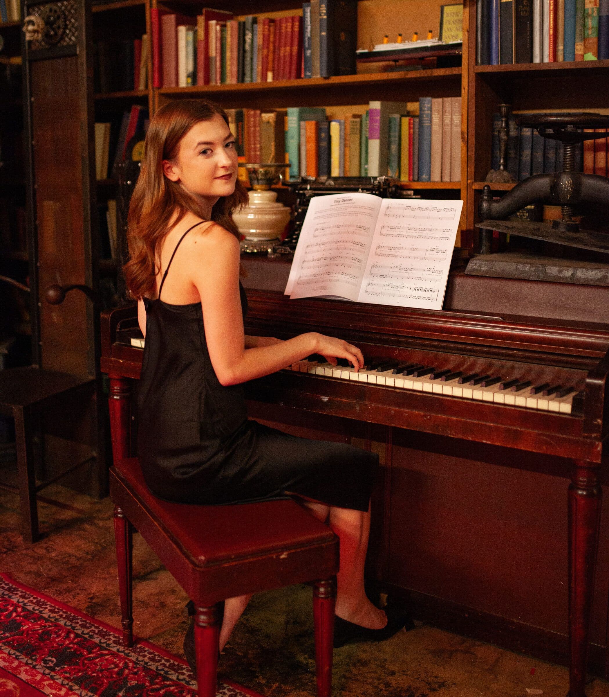 Thelma sits at an upright piano in a room walled with books, turning toward the camera, sheet music open in front of her.