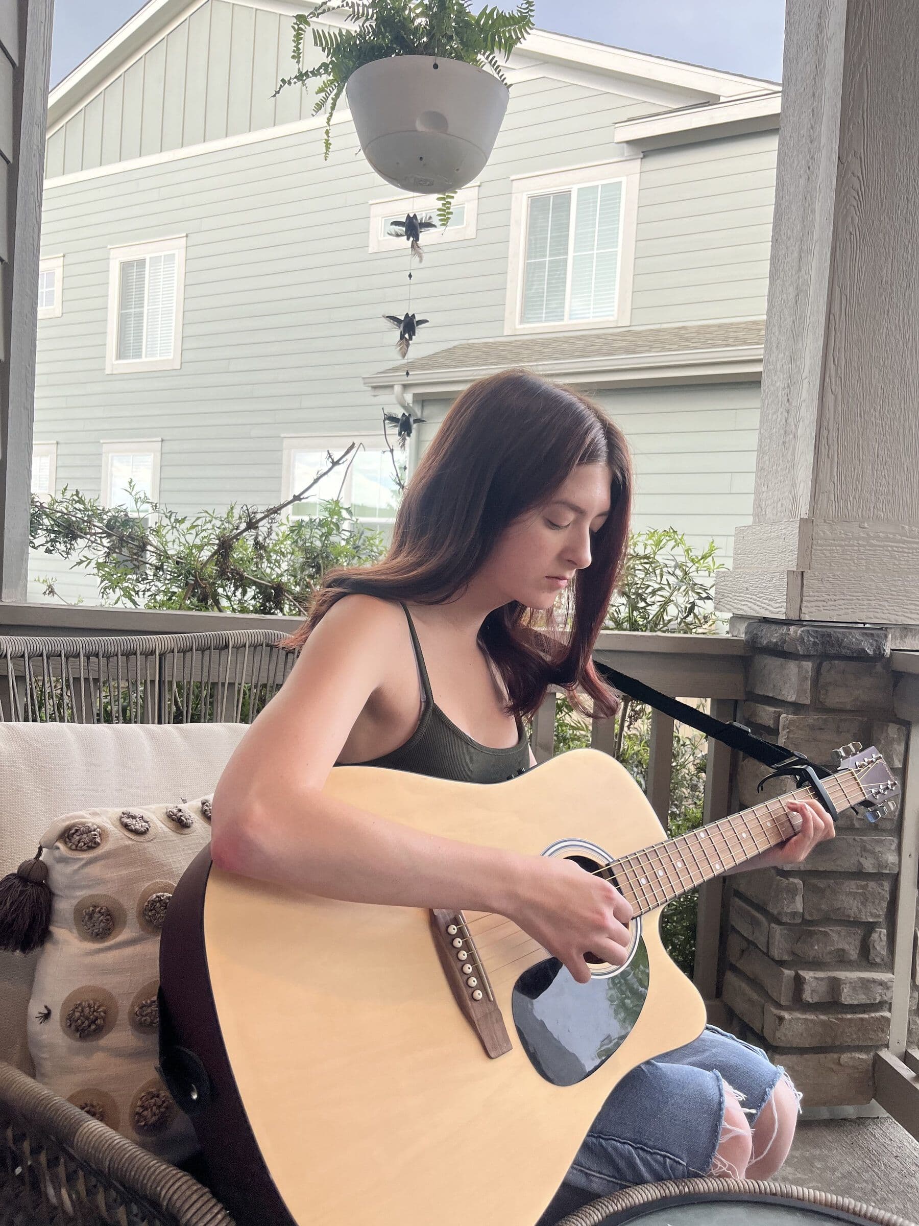 Thelma sits on a covered porch playing acoustic guitar, looking down at her hands. A hanging fern and a small bird mobile turn above her; a neighbouring house stands beyond the railing.
